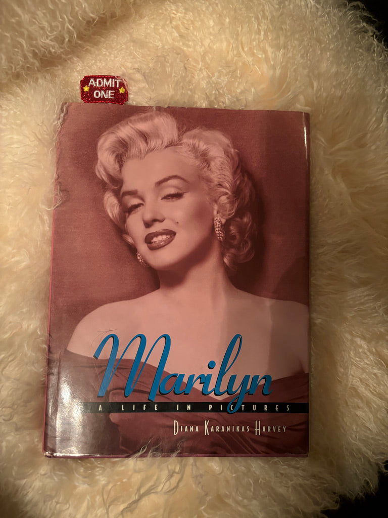 A picture of a book about Mariyln Monroe with a beautiful photo of her on the cover and a novelty Hollywood Movie themed bookmark in it, and the book is resting on a chair that has a furry texture to it.