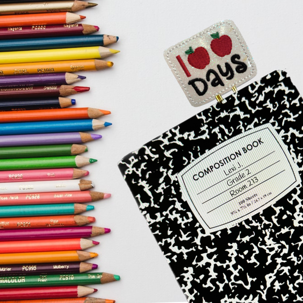 A Clip Chicks 100 Days novelty paper clip is shown sticking out of a black and white composition notebook, with a row of colored pencils laying beside the notebook. The novelty paper clip says 100 days with the zeros as the shape of red apples.