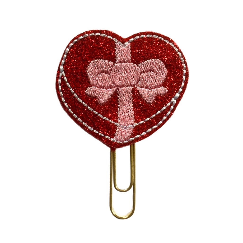 A novelty paper clip for Valentine's Day, with a feltie of red glittery vinyl shaped with a box of chocolates with a red bow embroidered on it. The feltie is attached to a gold toned paper clip making a playful journal accessory or bookmark.