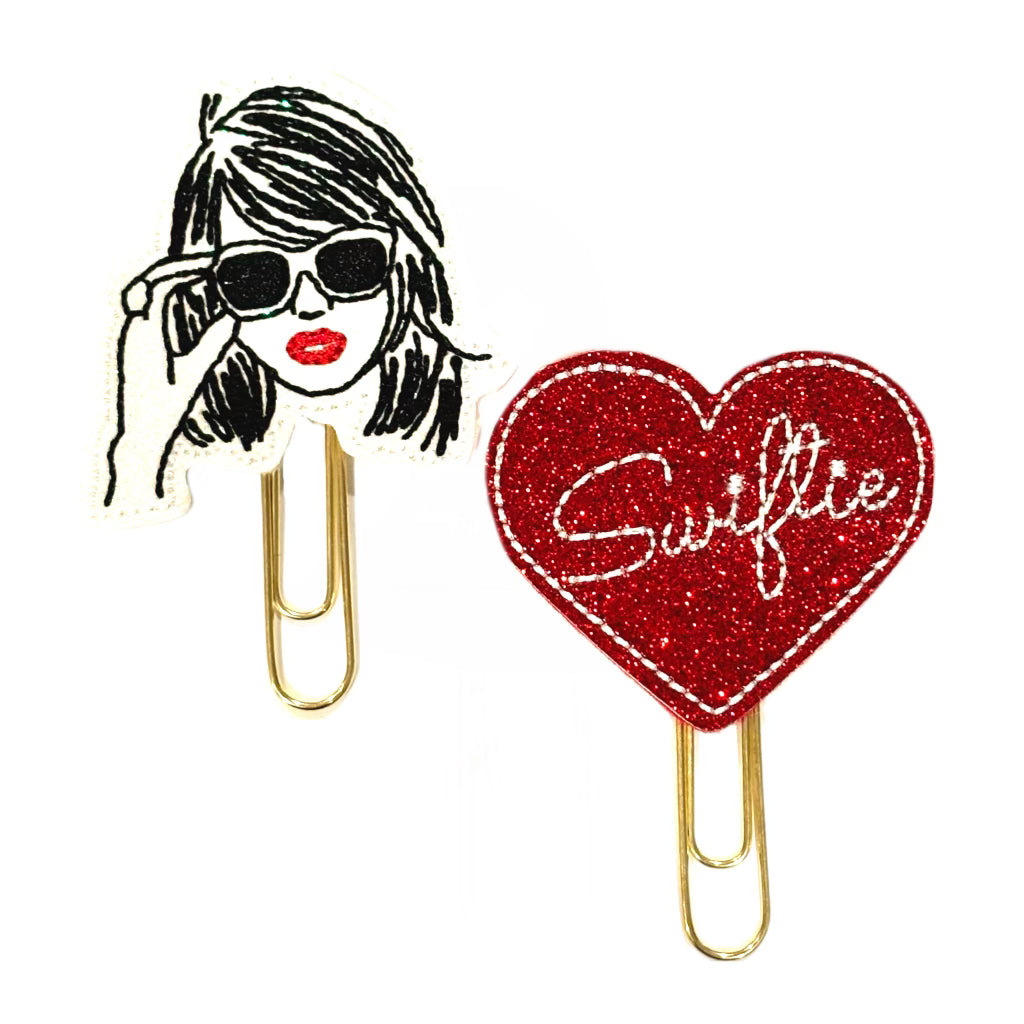 A set of two novelty paper clips, the paper clip is attached to an embroidered feltie. One feltie is embroidered to look like Taylor Swift's head with glasses on, the other is a red glittery heart with the word, Swiftie embroidered in white.