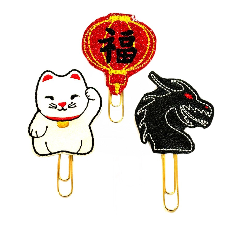 A trio of Lunar New Year novelty paper clips. Each feltie is attached to a gold paper clip, there is a lucky cat, a red lantern and a black dragon.