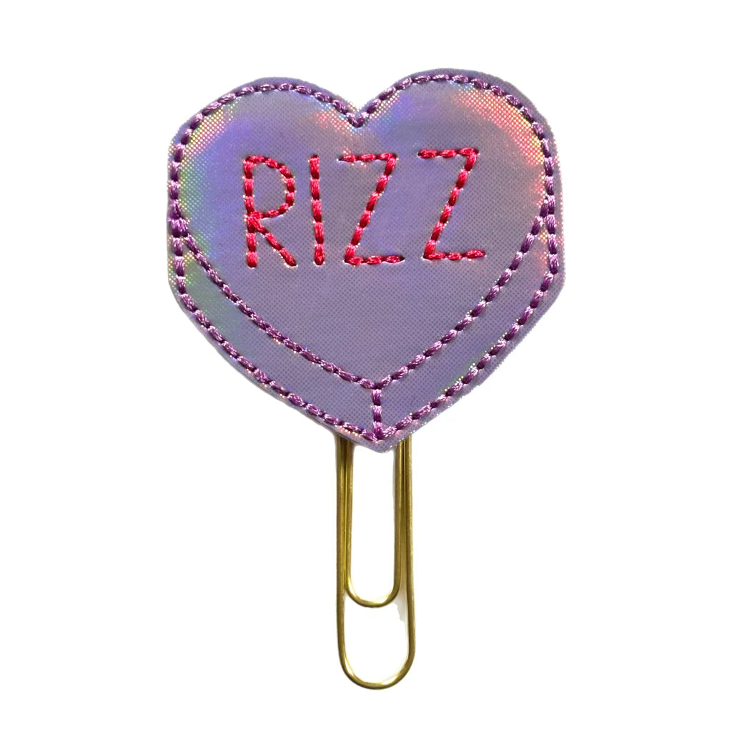 A feltie shaped with a lavender iridescent heart with the word, RIZZ, embroidered on it. the feltie is attached to a gold toned paper clip that can be used as a page marker in books, journals, planners, diaries, logs, magazines , etc. and also makes a great office accessory. 