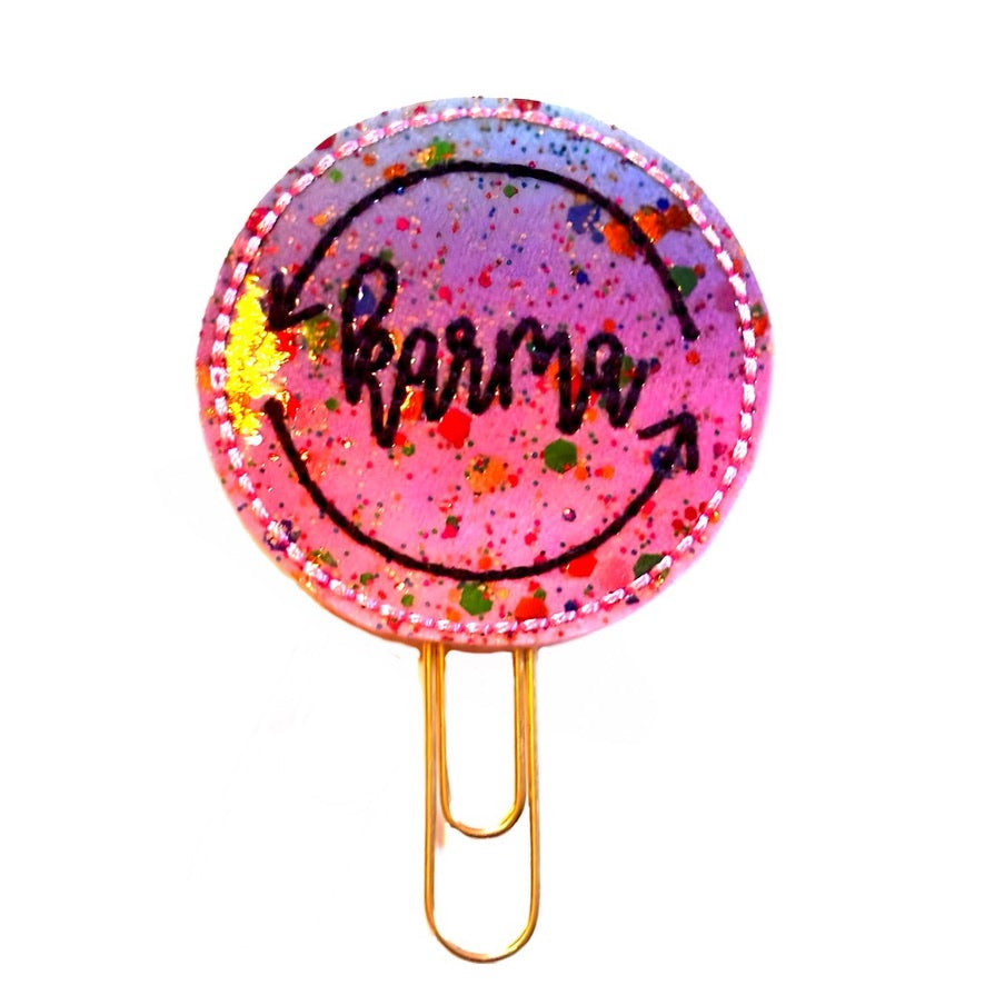 Shimmery ombre round feltie with the word, Karma, and circular arrows embroidered on it in black, then attached to a gold toned paper clip, for the Taylor Swift fan. 