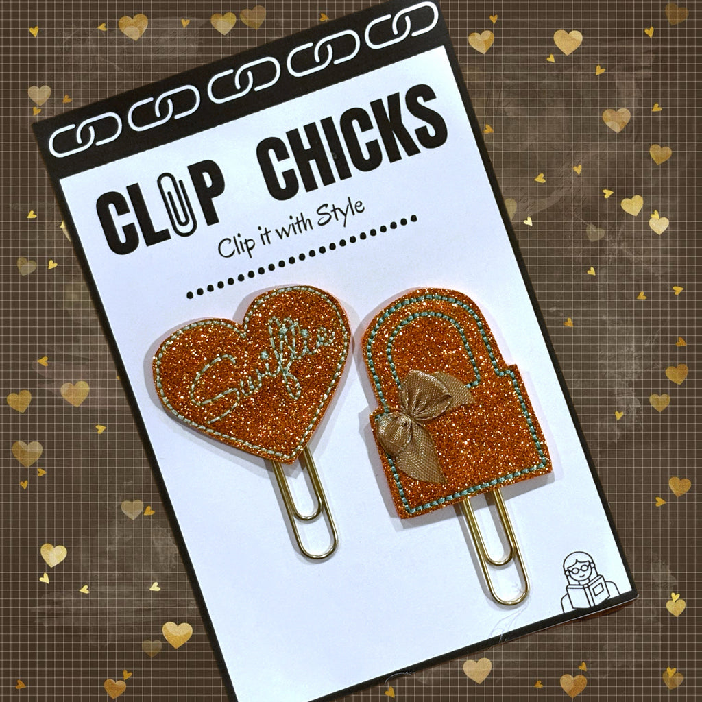 Set of orange shimmery Swiftie inspired paper clips from Clip Chicks LLC one is a heart and one a handbag shape.