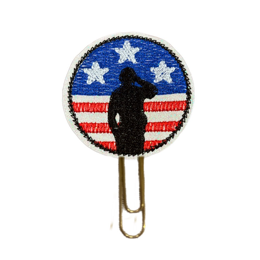 A novelty paper clip from Clip Chicks thats a round circle embroidered with stars, stripes and stars in red, white and blue and a saluting soldier. The embroidered feltie is then attached to a gold toned paper clip to use as a novelty bookmark, planner clip or as office decor .