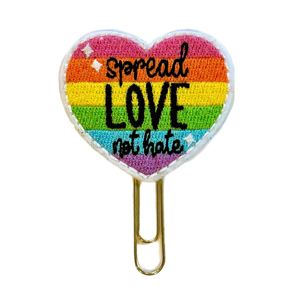 a heart shaped feltie with an embroidered rainbow and the words, Spread Love not Hate, on it. the feltie is attached to a gold toned paper clip and can be used as a bookmark, planner clip, journal accessory or paper organizer.