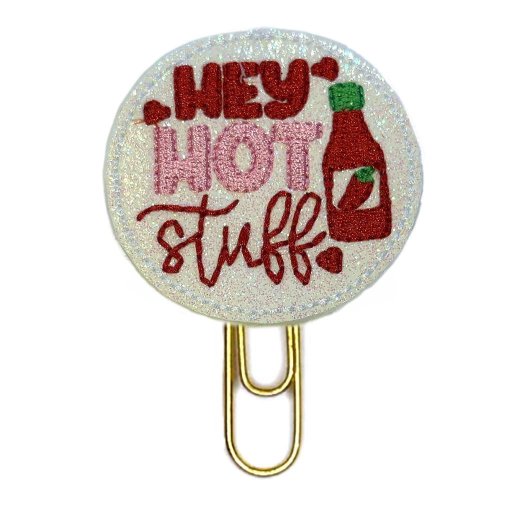 A novelty paper clip with a circular feltie made out of white glittery vinly with a red bottle of hot sauce and the words, Hey HOT Stuff, embroidered on it, then attached to a gold tonedd paper clip.