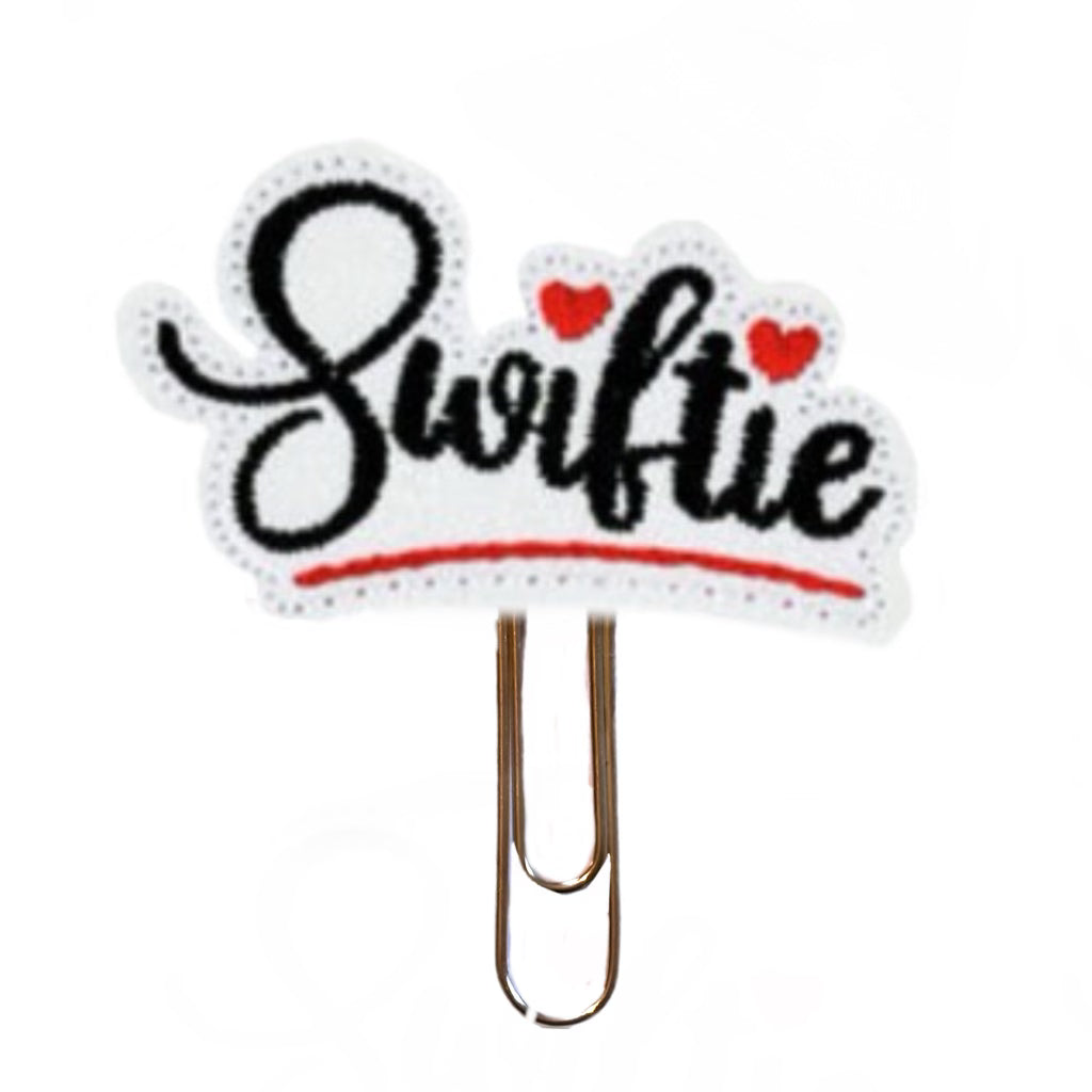 A novelty paper clip for Taylor Swift fans, the gold toned paper clip is attached to a glittery white vinyl feltie that is embroidered with the word, Swiftie, on it as well as little red hearts.