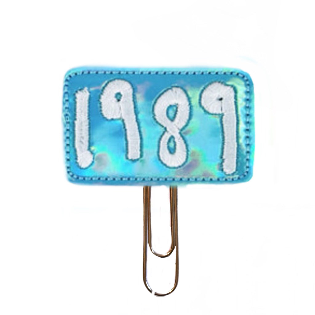 1989 embroidered on a rectangular shiny blue vinyl feltie and attached to a gold toned paper clip to use as a bookmark or page markers or paper organizer.