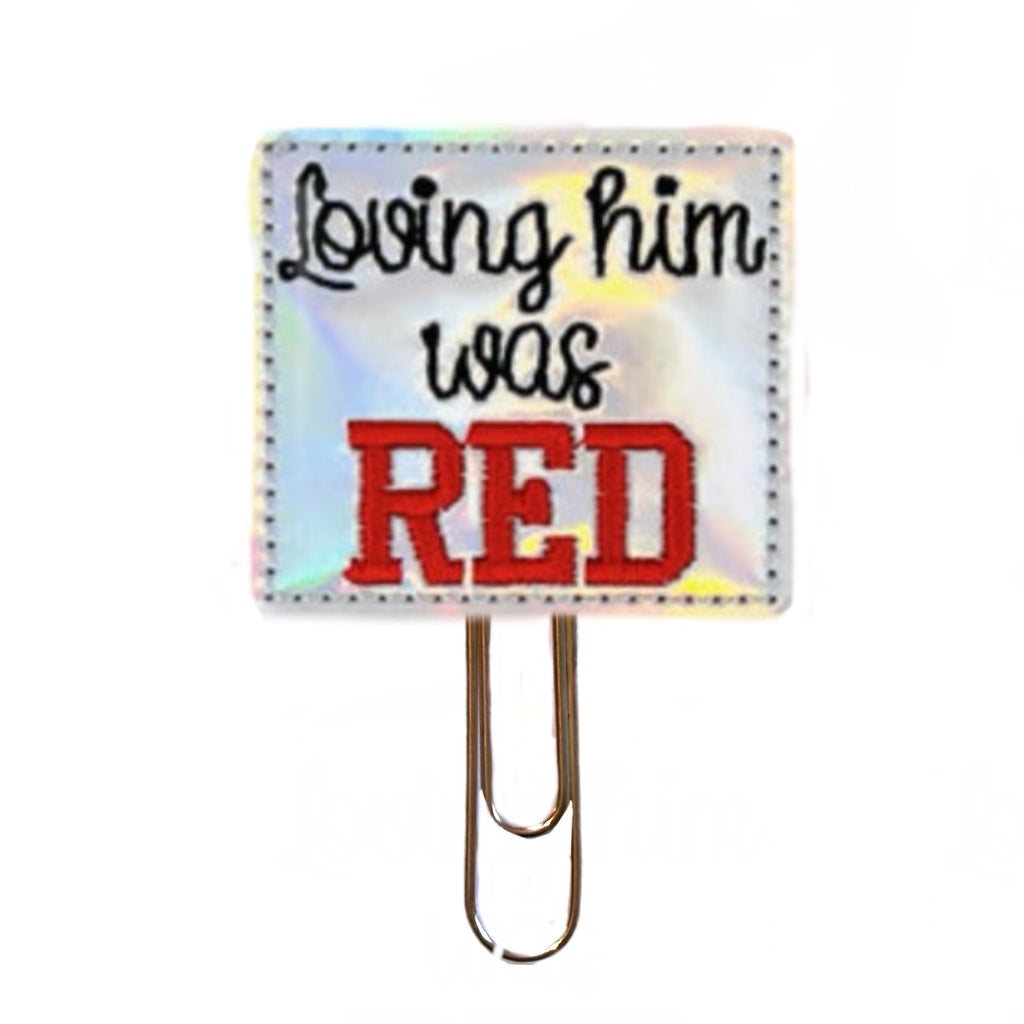 A shiny silver feltie made from vinyl and felt, with the words, Loving him was RED, embroidered on it, then attached to a gold toned paper clip.