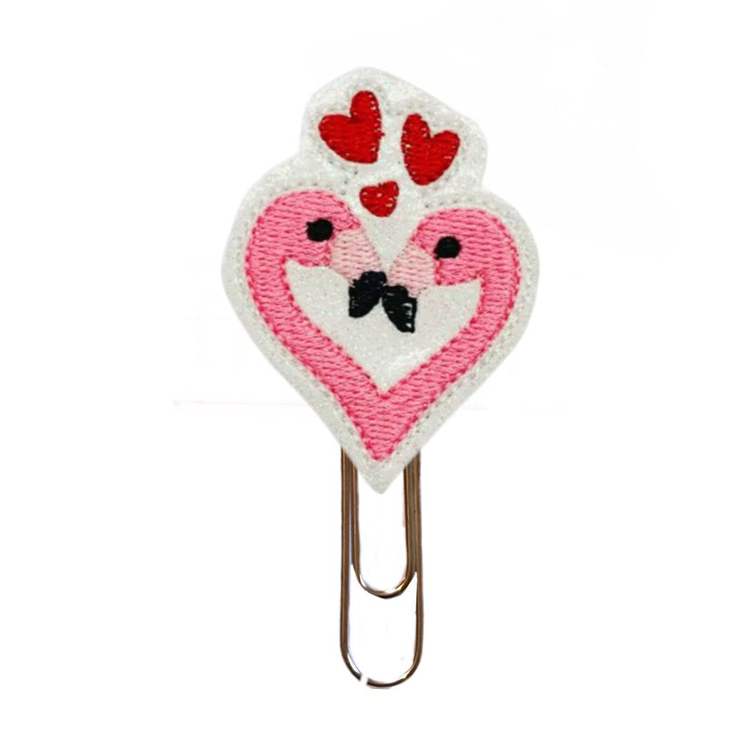 A novelty paper clip with a feltie shaped like a heart with two embroidered flamingo heads forming a heart with three hearts floating above them. Feltie is attached to a gold toned paper clip.