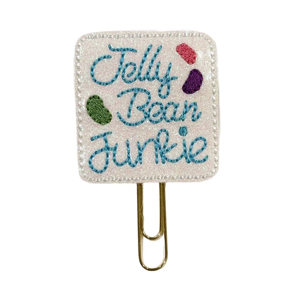 A Clip Chicks novelty paper clip attached to a white shimmery feltie where Jelly bean Junkie and three colorful jelly beans are embroidered onto the feltie,