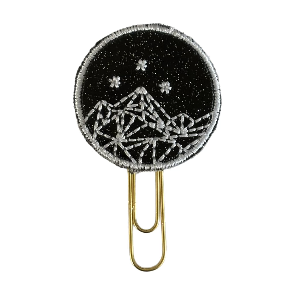 A picture of Cliuo Chicks ACOTAR insipired novelty paper clip. The gold paperclip is attached to a black shimmery vinyl circle with white embroidery on it, making a mountainscape with three stars in the sky above the maountains.