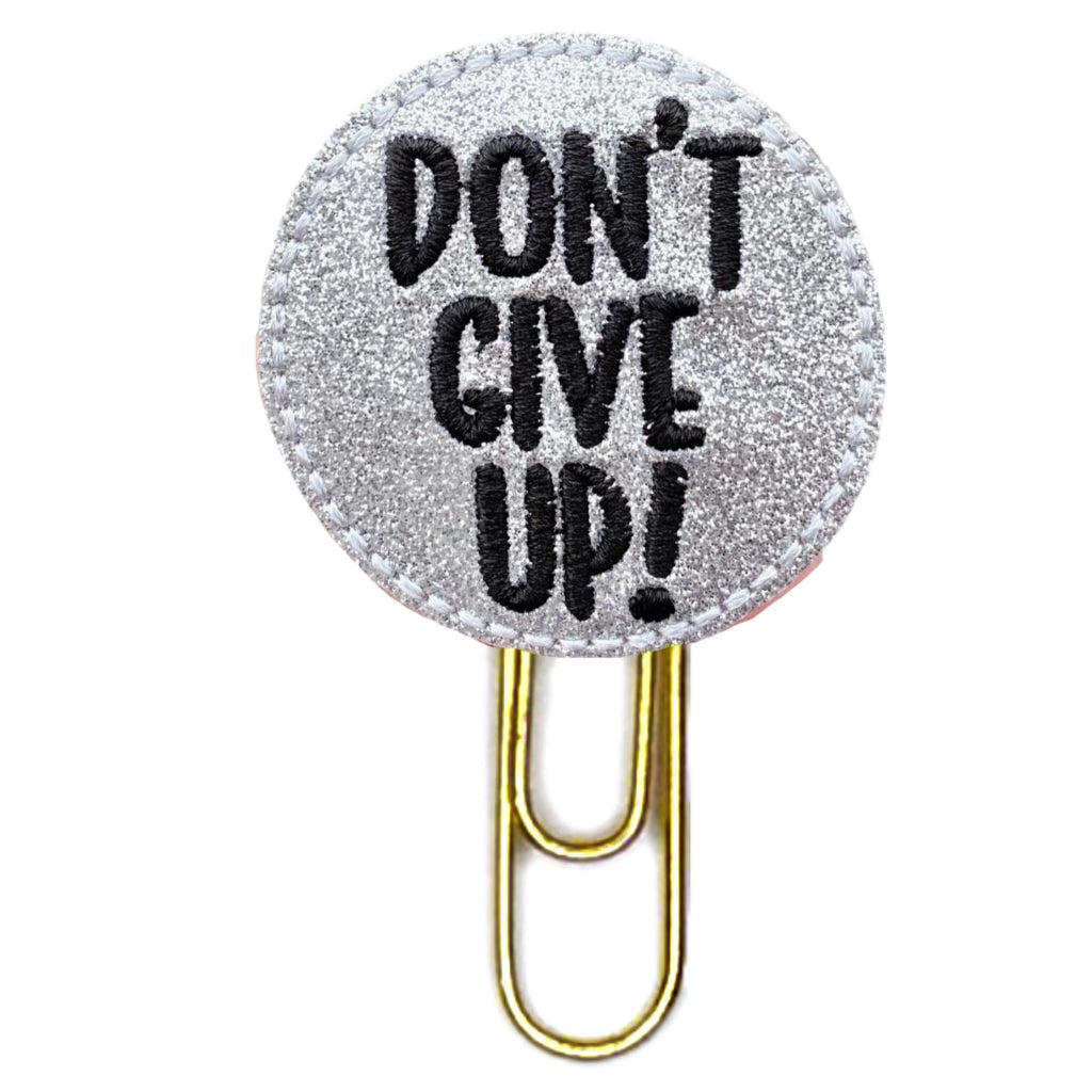 Don;t Give Up is embroidered on a two inch circle made of shiny silver vinyl, and the thread is black, then attached to a gold-toned paper clip.