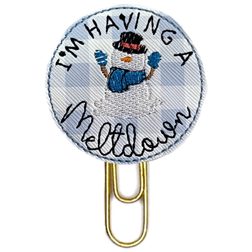 Snowman and Having a meltdown are embroidered on a blue and white plaid circular piece of vinyl then attached to a gold toned paper clip.