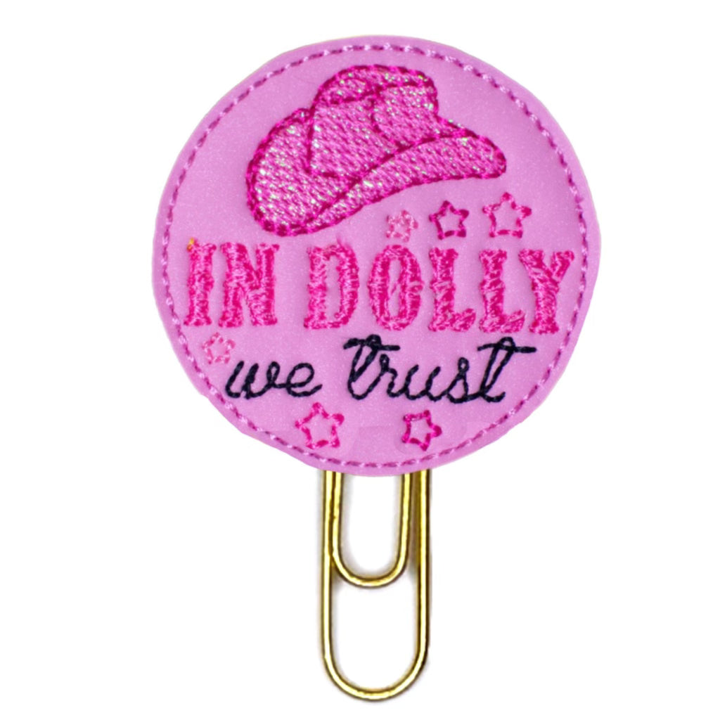 Pink round feltie that reads In Dolly We Trust and has a pink cowboy hat, and is attached to a gold-toned paper clip.