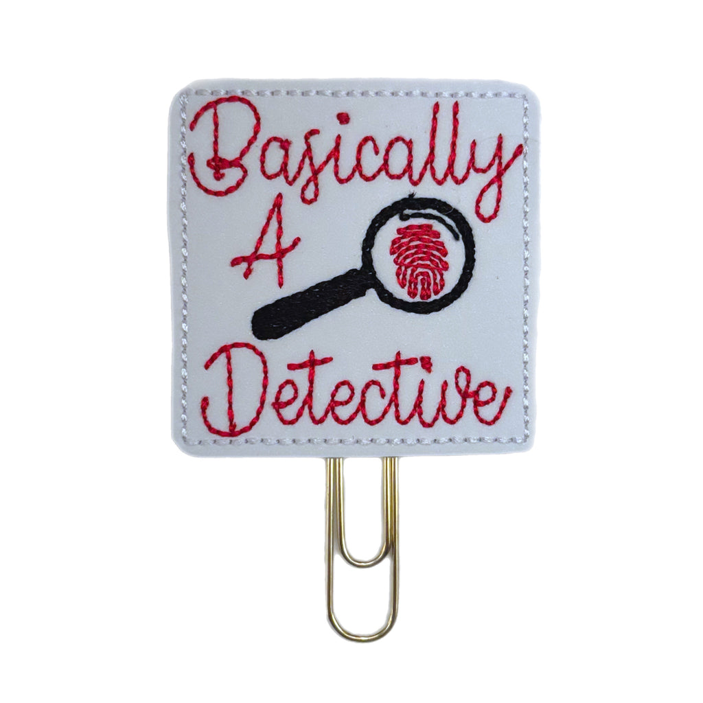 A white 2 inch square feltie with the words, Basically A Detective embroidered on it in red, with a black magnifying glass and a fingerprint. The feltie is attached to a gold tone paper clip and used as a bookmark or page saver.